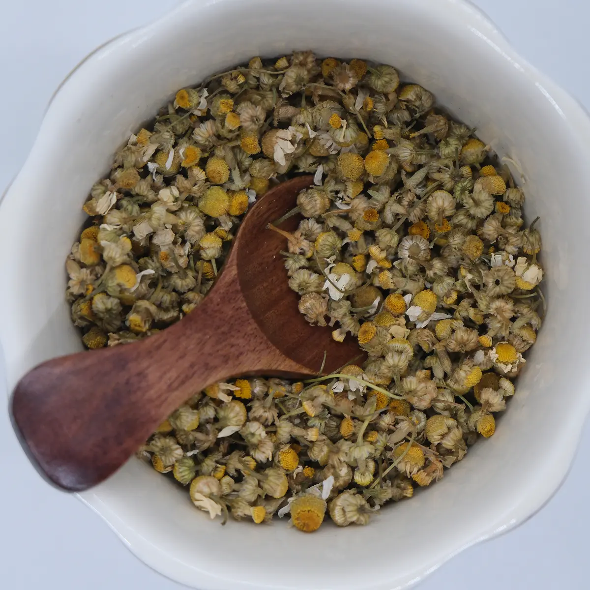 Whole dried chamomile flowers in a white bowl with a wooden scoop