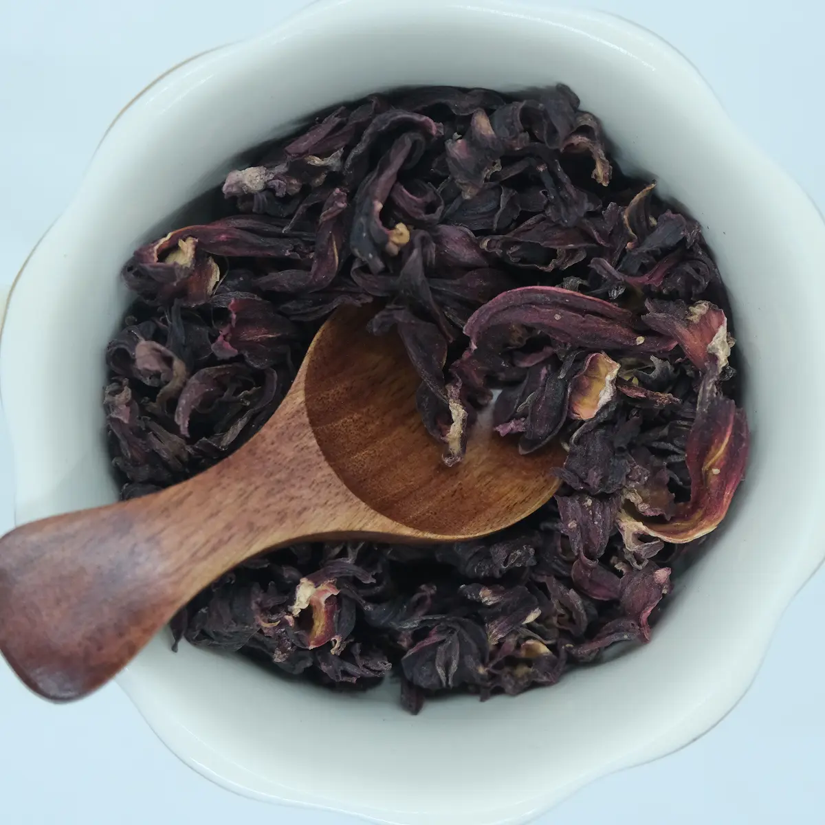 Deep red dried hibiscus petals in a white bowl with a wooden scoop