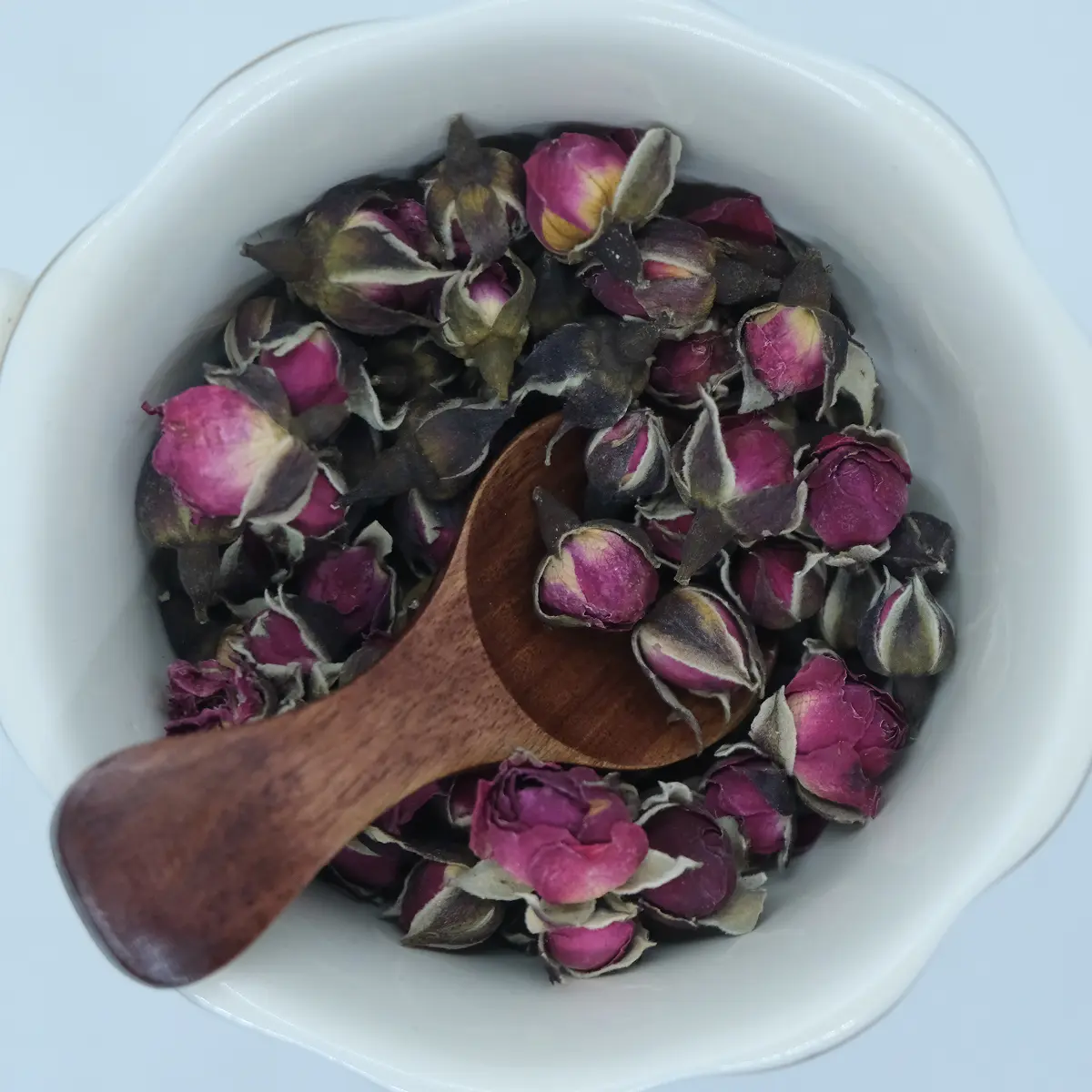 Whole dried pink rose buds in a white bowl with a wooden scoop